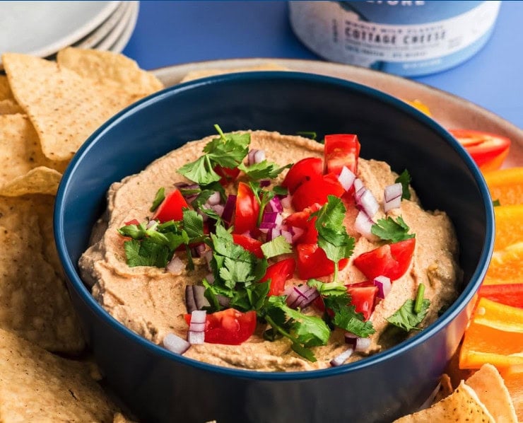 A blue bowl of hummus topped with chopped tomatoes, red onions, and cilantro, surrounded by tortilla chips and sliced bell peppers—perfect alongside other cottage cheese breakfast recipes for a colorful spread.