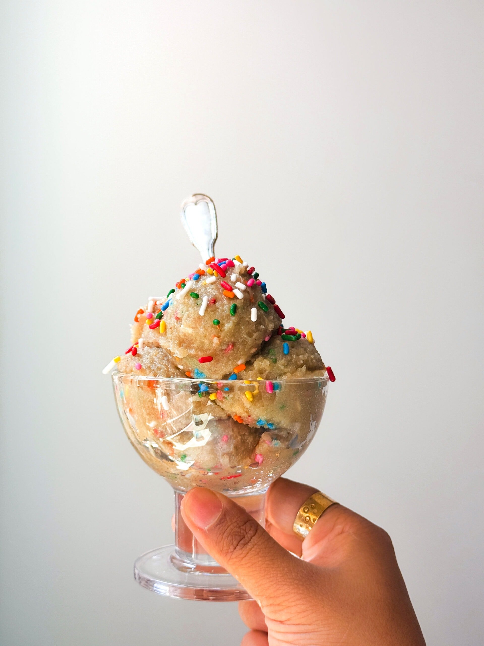 A hand holding a glass bowl of cottage cheese ice cream topped with colorful sprinkles and a small plastic spoon, against a plain background—an inviting high-protein treat.