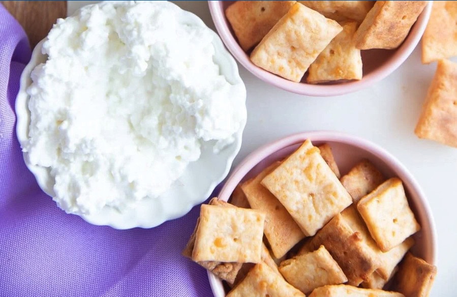 A small white bowl filled with cottage cheese sits on a light surface next to two pink bowls filled with golden-brown, square-shaped crackers. A lavender cloth is partially visible in the corner.