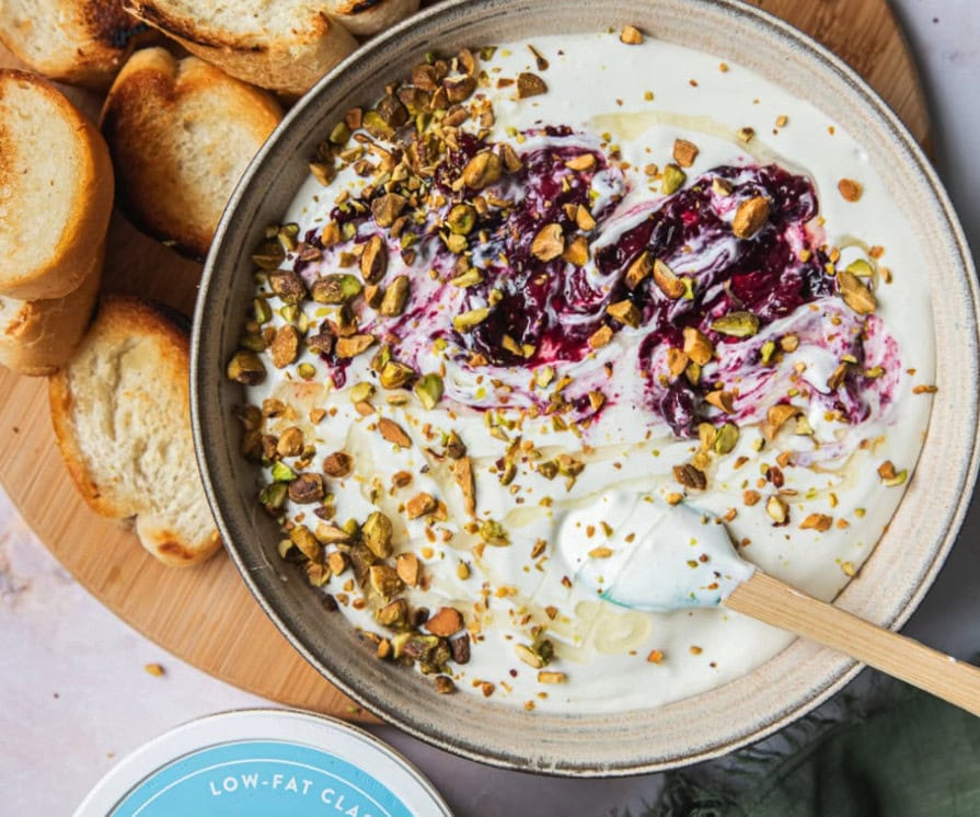 A bowl of yogurt topped with crushed pistachios and berry sauce, with a spatula resting inside—perfect for summer entertaining. Slices of toasted bread are arranged on the side for a delightful high-protein snack.