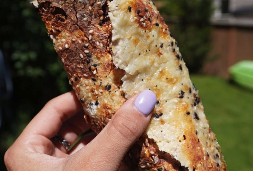 A hand with light purple nail polish holds a piece of crusty, seeded bread topped with melted cheese, with a blurred green lawn and fence in the background.
