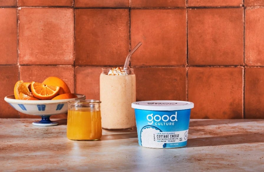 A container of Good Culture low-fat classic cottage cheese sits on a countertop next to a jar of honey, a glass of creamy smoothie topped with granola and a straw, and a bowl of sliced oranges, with a warm brown tiled wall in the background.