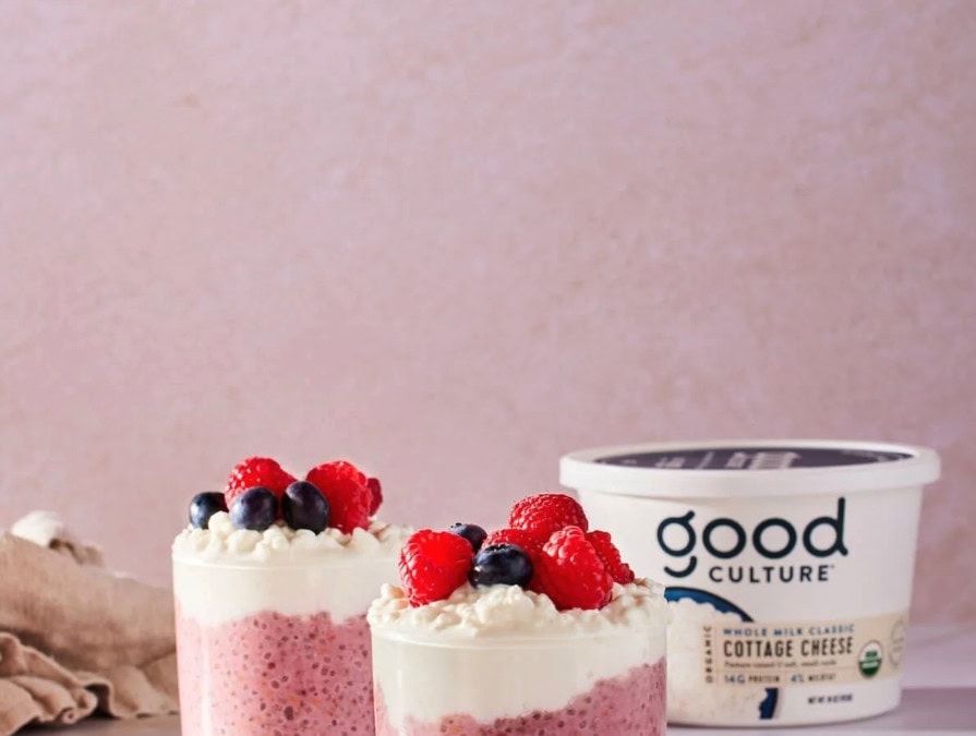 Two glass cups filled with layers of pink chia pudding and creamy cottage cheese, topped with fresh raspberries and blueberries, sit in front of a container of Good Culture whole milk classic cottage cheese against a soft pink background.