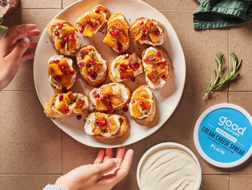 Toasted baguette slices topped with roasted butternut squash, pomegranate seeds, and Good Culture cream cheese spread, arranged on a plate beside a container of Good Culture Plain Cream Cheese Spread.