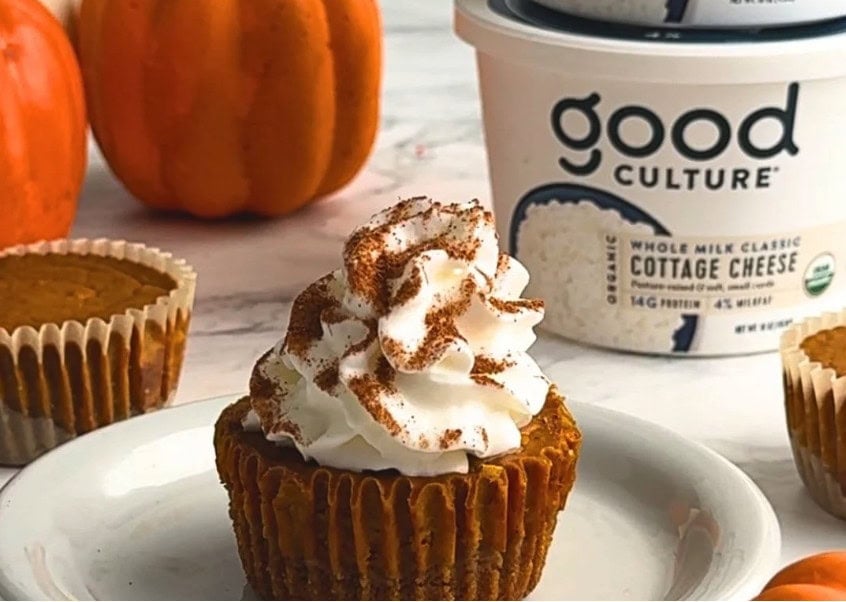 Mini pumpkin cheesecakes topped with whipped cream and cinnamon, displayed with Good Culture cottage cheese containers and small pumpkins in the background.